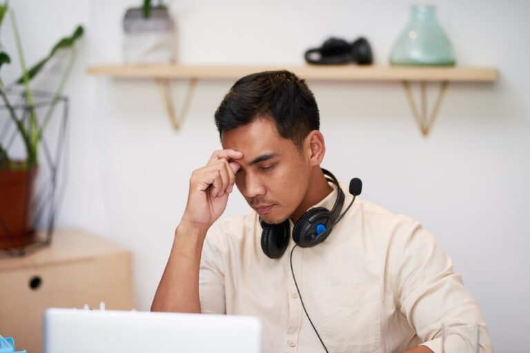 Upset looking man with hand on forehead, eyes closed, in front of laptop, headphones around neck